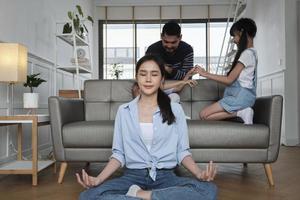 une jeune mère thaïlandaise asiatique est assise sur le sol du salon, médite et pratique le yoga pour la santé et le bien-être lorsque papa et les enfants jouent ensemble sur un canapé, mode de vie domestique heureux le week-end en famille. photo