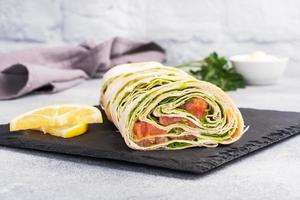 rouleaux de pain pita fin et de saumon salé rouge avec des feuilles de laitue sur un support en ardoise, fond de béton gris. espace de copie. photo