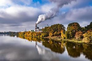 tuyaux de la scierie de l'usine de menuiserie près de la rivière avec des arbres jaunes rouges d'automne. notion de pollution atmosphérique. paysage industriel pollution environnementale déchets de centrale thermique photo