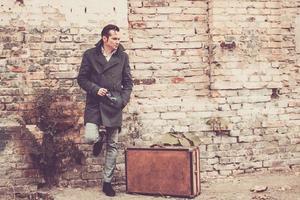 portrait d'un homme réfléchi de style vintage avec une valise en cuir appuyée sur un mur de briques. photo
