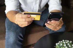 homme tenant une carte de crédit et utilisant un smartphone à la maison, homme d'affaires faisant des achats en ligne, commerce électronique, services bancaires par Internet, dépenser de l'argent, travailler à domicile, s'asseoir sur le canapé dans le salon à la maison. photo