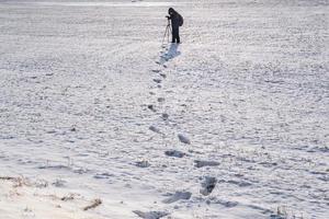 photographe avec un trépied dans un champ enneigé prend des photos de paysage d'hiver, des empreintes de pas dans la neige