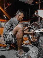 un vieil homme en short court s'efforce de changer le pneu arrière de la moto la nuit dans son atelier. photo prise la nuit à l'extérieur dans le village de luhu, gorontalo le 21 août 2022