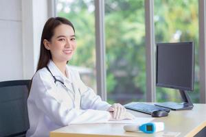 asiatique belle jeune femme souriante médecin assis au bureau à l'hôpital. sur la table a un papier et un ordinateur. photo