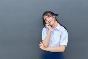jolie lycéenne asiatique en uniforme scolaire avec des sourires en toute confiance pendant qu'elle regarde la caméra joyeusement avec du gris en arrière-plan. photo