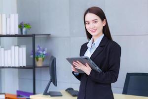 une travailleuse asiatique en costume noir tient un ipad ou une tablette dans ses mains et sourit dans la salle de bureau. photo
