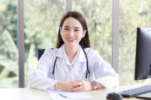 belle femme médecin asiatique assis sourire vêtu d'une robe blanche et d'un stéthoscope en attendant un patient au concept de soins de santé hospitaliers. photo