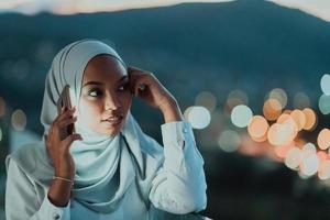 jeune femme musulmane portant un voile d'écharpe dans la rue de la ville urbaine la nuit envoyant des SMS sur un smartphone avec la lumière de la ville bokeh en arrière-plan. photo