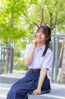 jolie étudiante asiatique du secondaire dans l'uniforme scolaire s'assoit en toute confiance et sourit joyeusement pendant qu'elle regarde la caméra dans un parc de la ville. photo