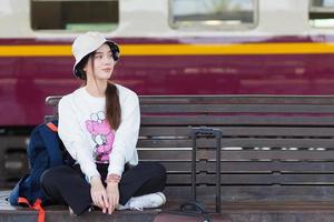 une femme asiatique en manches longues blanches et un chapeau est assis sur une chaise avec son sac sur la plate-forme du train dans la distanciation sociale comme un nouveau mode de vie normal. photo