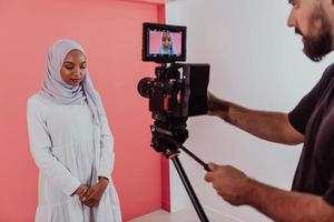 vidéaste en studio numérique enregistrant une vidéo sur une caméra professionnelle en tirant sur une femme musulmane portant une écharpe hijab sur fond rose en plastique. photo