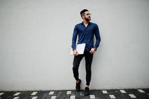 un entrepreneur du moyen-orient porte une chemise bleue, des lunettes, tient un ordinateur portable contre un mur blanc. photo