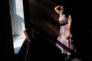 jeu avec les ombres. portrait de mode d'une femme afro-américaine en robe longue et lunettes à l'intérieur. photo