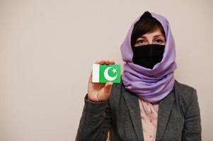 portrait d'une jeune femme musulmane portant des vêtements de cérémonie, protégez le masque facial et le foulard hijab, tenez la carte du drapeau pakistanais sur fond isolé. concept de pays coronavirus. photo