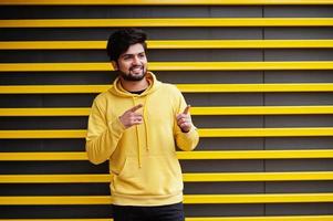 jeune homme indien hipster urbain dans un sweat-shirt jaune à la mode. un gars sud-asiatique cool porte un sweat à capuche sur fond rayé montrant les doigts pour libérer de l'espace. photo