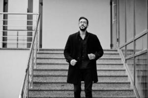 un entrepreneur du moyen-orient porte un manteau noir et une chemise bleue, des lunettes contre un immeuble de bureaux marchant dans les escaliers. photo