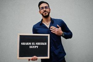 l'homme arabe porte une chemise bleue et des lunettes tient le tableau avec l'inscription de sharm el sheikh egypte. plus grandes villes du concept du monde islamique. photo