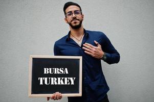 l'homme arabe porte une chemise bleue et des lunettes tiennent le tableau avec l'inscription bursa turquie. plus grandes villes du concept du monde islamique. photo