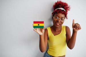 femme africaine aux cheveux afro, porter un maillot jaune et des lunettes, tenir le drapeau du ghana isolé sur fond blanc, montrer le pouce vers le haut. photo