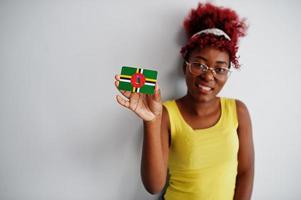 femme afro-américaine aux cheveux afro, porter un singulet jaune et des lunettes, tenir le drapeau de la dominique isolé sur fond blanc. photo