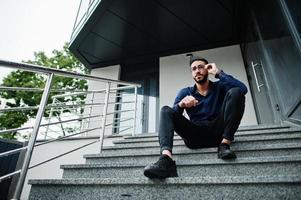 un entrepreneur du moyen-orient porte une chemise bleue, des lunettes contre un immeuble de bureaux assis dans les escaliers. photo