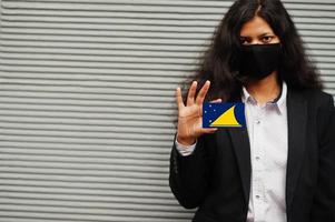 une femme asiatique en tenue de soirée et un masque de protection noir tiennent le drapeau tokelau à portée de main sur fond gris. coronavirus au concept de pays. photo