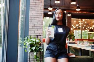 femme afro-américaine féministe à la mode vêtue d'un t-shirt et d'un short noirs, posée au restaurant avec un verre de limonade. photo