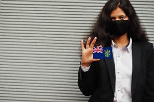 une femme asiatique en tenue de soirée et un masque de protection noir tiennent le drapeau des îles pitcairn à portée de main sur fond gris. coronavirus au concept de pays. photo