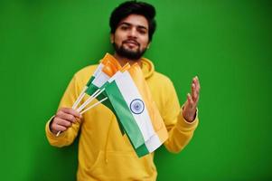 un jeune indien hipster urbain dans un sweat-shirt jaune à la mode tient des drapeaux de l'inde dans les mains. un gars sud-asiatique cool porte un sweat à capuche isolé sur fond vert. photo