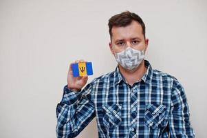 homme en chemise à carreaux montre la carte du drapeau de la barbade à la main, porte un masque de protection isolé sur fond blanc. concept de coronavirus des pays américains. photo