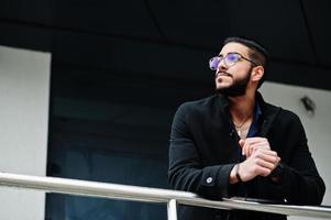 un entrepreneur du moyen-orient porte un manteau noir et une chemise bleue, des lunettes contre un immeuble de bureaux. photo