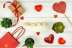 composition romantique créative de la saint-valentin avec coeurs rouges, ruban de satin, sucette, boîte-cadeau et sac en papier sur fond blanc. maquette avec des cubes en bois vides pour le texte. photo