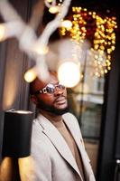 un homme afro-américain élégant porte une veste beige avec des lunettes de soleil pose contre la maison avec une guirlande dorée. photo