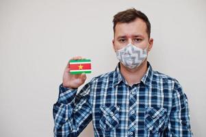 homme en chemise à carreaux montre la carte du drapeau du suriname à la main, porte un masque de protection isolé sur fond blanc. concept de coronavirus des pays américains. photo