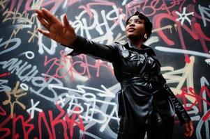 belle femme afro-américaine à la mode posant dans une veste et un pantalon en cuir noir dans la rue contre le mur de graffitis. photo