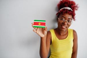 femme afro-américaine aux cheveux afro, porter un singulet jaune et des lunettes, tenir le drapeau du suriname isolé sur fond blanc. photo