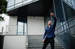 un entrepreneur du moyen-orient porte une chemise bleue, des lunettes avec un ordinateur portable contre un immeuble de bureaux. photo