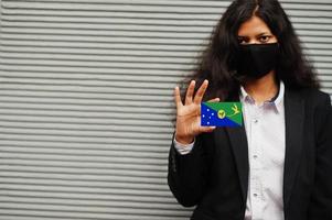 femme asiatique en tenue de soirée et masque de protection noir tenir le drapeau de l'île de noël à portée de main sur fond gris. coronavirus au concept de pays. photo