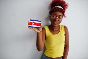 femme afro-américaine aux cheveux afro, porter un singulet jaune et des lunettes, tenir le drapeau du costa rica isolé sur fond blanc. photo