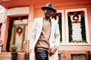 un homme afro-américain élégant porte une veste beige et un chapeau noir avec des lunettes de soleil pose contre la maison avec des décorations et une couronne du nouvel an. photo