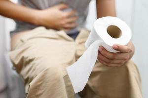 l'homme souffre de diarrhée main tenir le rouleau de papier de soie devant la cuvette des toilettes. constipation dans la salle de bain. traitement des douleurs à l'estomac et de l'hygiène, soins de santé photo