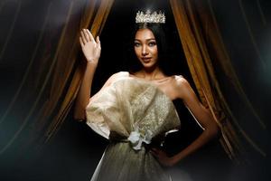 silhouette de concours de concours de beauté miss femme avec une couronne de diamants scintillants avec une peau bronzée belle robe de soirée de maquillage sur scène avec rideau d'éclairage et fond sombre photo