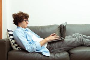 un jeune homme caucasien travaille à domicile sur un ordinateur portable et se connecte en ligne au bureau à l'intérieur pour un lieu de travail Internet à distance. jeune homme adulte est un travail indépendant intelligent sur un ordinateur portable allongé sur un canapé-lit, espace de copie photo