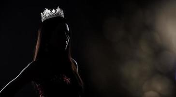 portrait du concours de beauté de miss pageant en robe de soirée à sequins robe longue avec couronne de diamants scintillants, silhouette exposition discrète avec rideau, éclairage de studio fond sombre dramatique photo