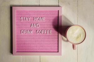 vue de dessus de la tasse rose avec cappuccino et qoute restez à la maison et buvez du café. campagne d'auto-isolement et de quarantaine pour vous protéger pendant la pandémie de coronavirus. photo
