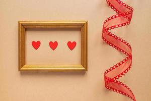 concept romantique de la saint-valentin ou du mariage. ruban décoratif torsadé et cadre photo doré avec coeurs rouges sur fond beige.