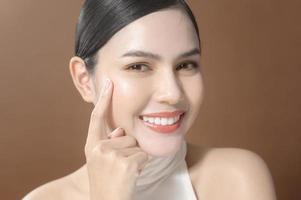 une jeune femme avec un beau visage souriant, touchant son visage sur fond marron, concept de soins de la peau beauté photo