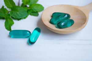 capsules bleues dans une cuillère en bois avec des feuilles de menthe sur fond de bois blanc. photo