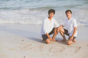 jeune couple asiatique gay souriant dessin romantique mot texte amour ensemble sur le sable en vacances, homosexuel heureux et amusant avec amour assis sur le sable à la plage en voyage d'été, concept juridique lgbt. photo