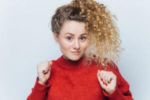 portrait d'une belle femme aux cheveux bouclés, lève les poings, vêtue d'un pull rouge décontracté, isolée sur fond blanc, pose en studio. les gens, la beauté, les expressions faciales et le concept de langage corporel photo